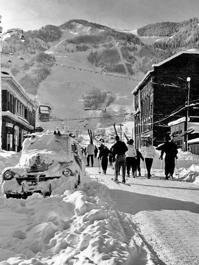 Vintage skiers walk up mill street toward the slopes